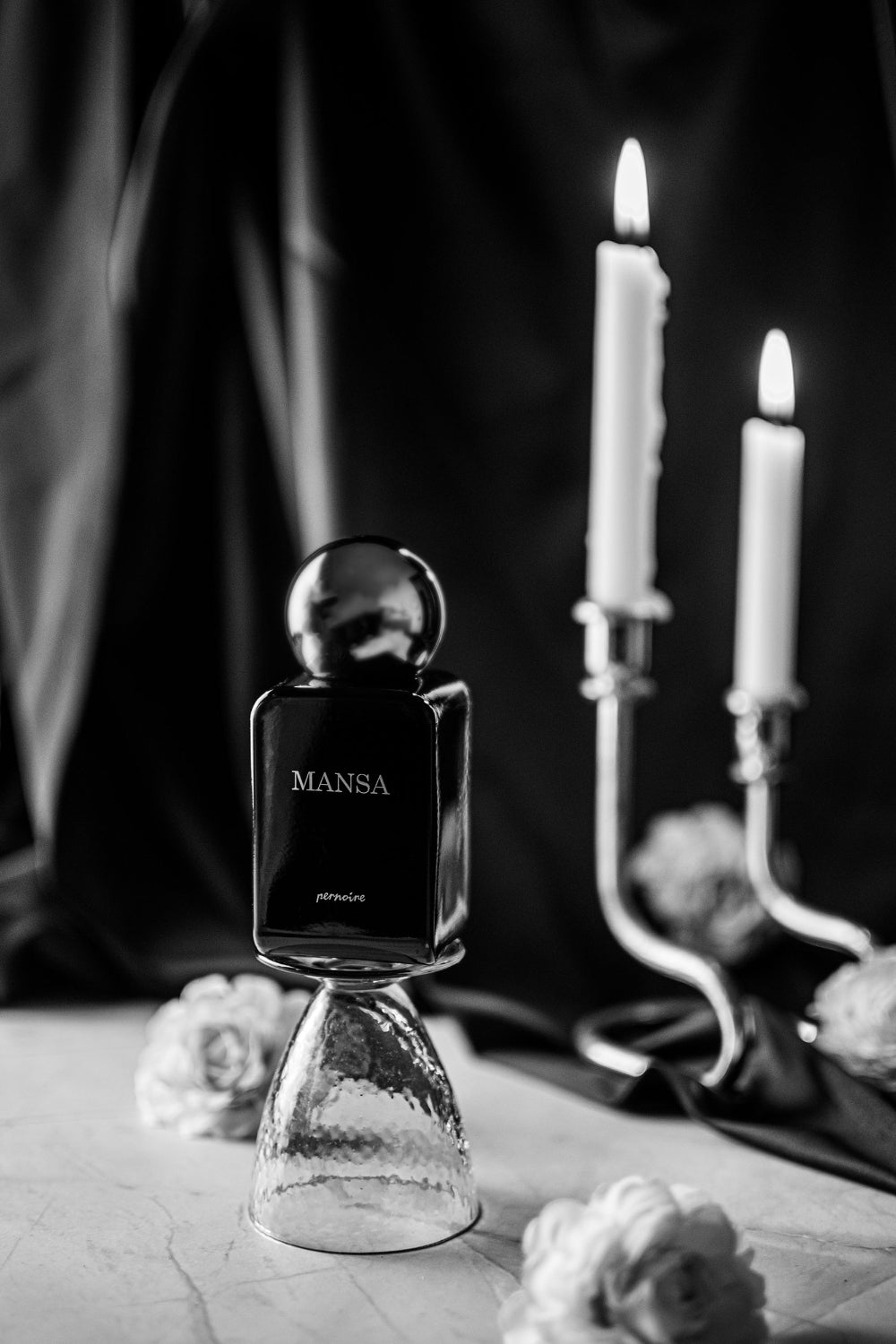 Originaler MANSA-Flakon auf einem umgedrehten Glas neben Rosen und zwei brennenden Kerzen.