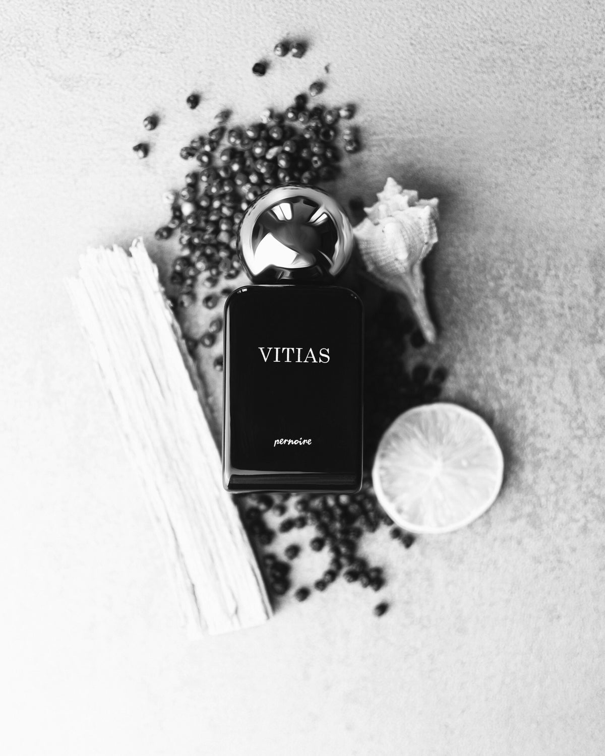 Black VITIAS bottle on pale sand with a shell, a citrus slice, peppercorns and weathered wood.