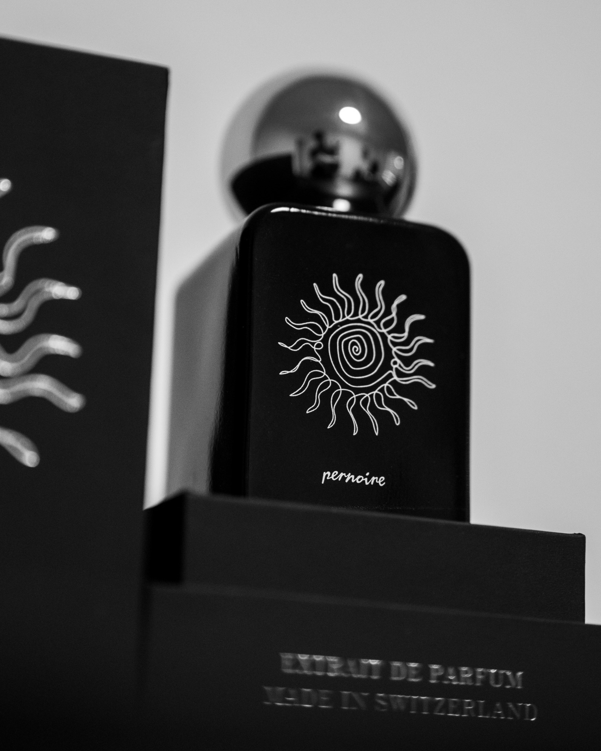 Schwarzer TIERRA-Flakon mit spiralförmiger Sonnenzeichnung in geöffneter schwarzer Verpackung vor heller Wand.