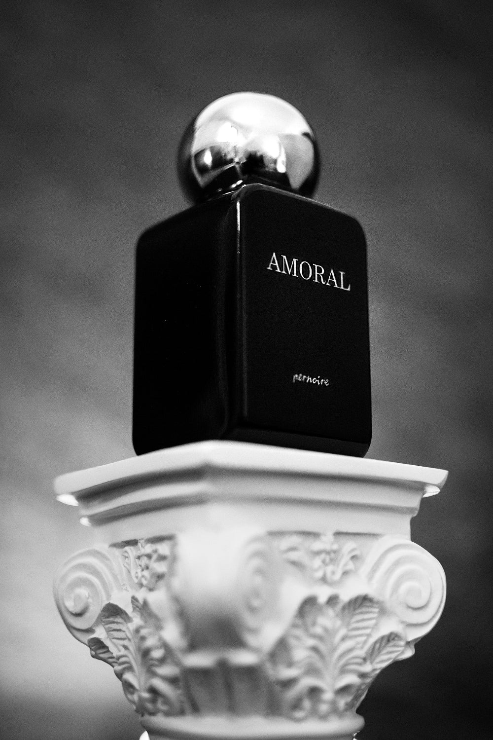 Black AMORAL perfume bottle by pernoire on an ornate white column, with the fragrance name facing forward.