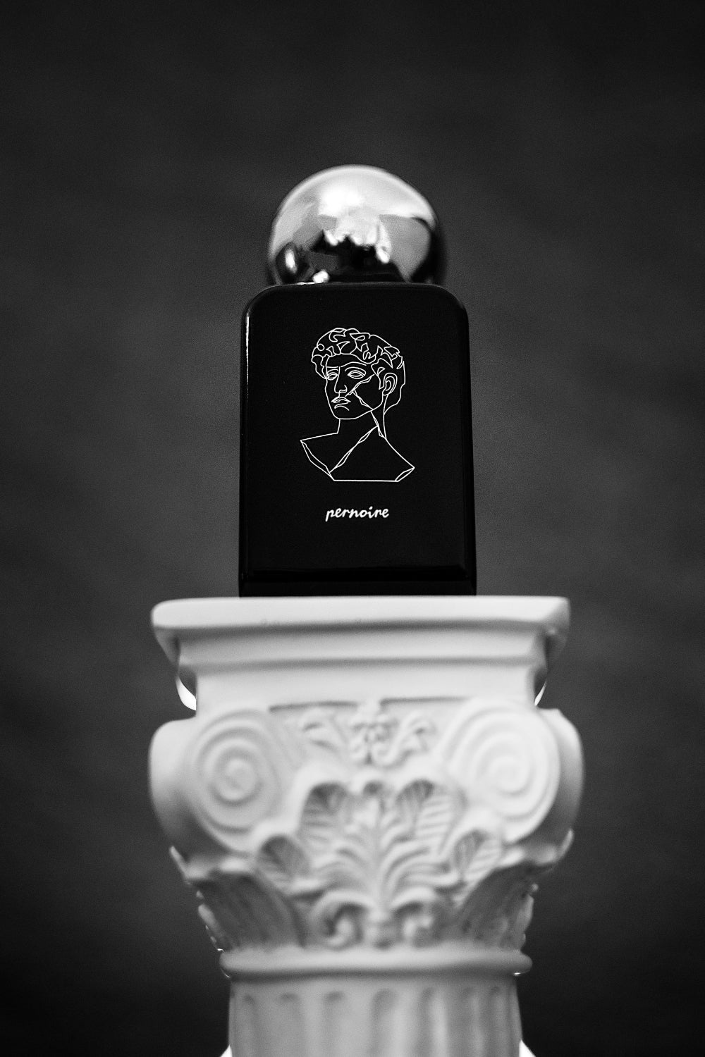Originaler AMORAL-Flakon mit Gesichtssymbol und pernoire-Logo auf einer verzierten weissen Säule.