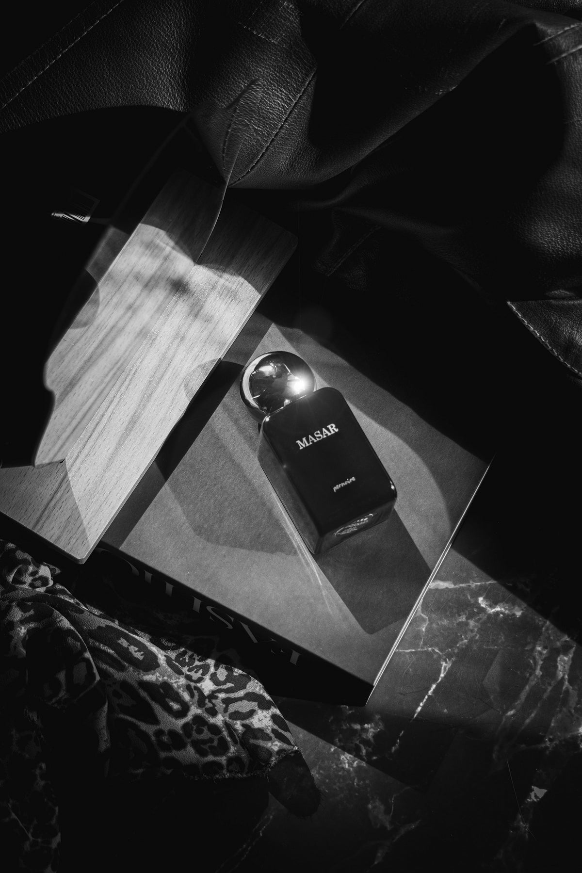 Black MASAR bottle on an open magazine beside dark leather and patterned fabric.