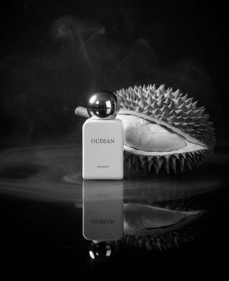 Weisser OUDIAN-Flakon neben geöffneter Durian auf dunklem, spiegelndem Tisch mit Rauchschleiern.