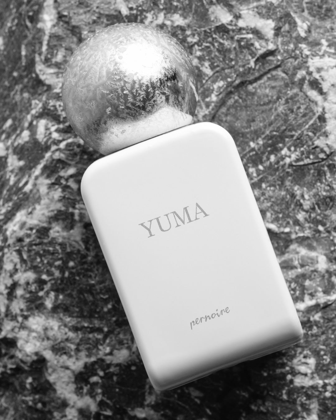 Aufnahme des weissen YUMA-Flakons auf strukturiertem Stein, mit Duftnamen und bereifter Kappe.