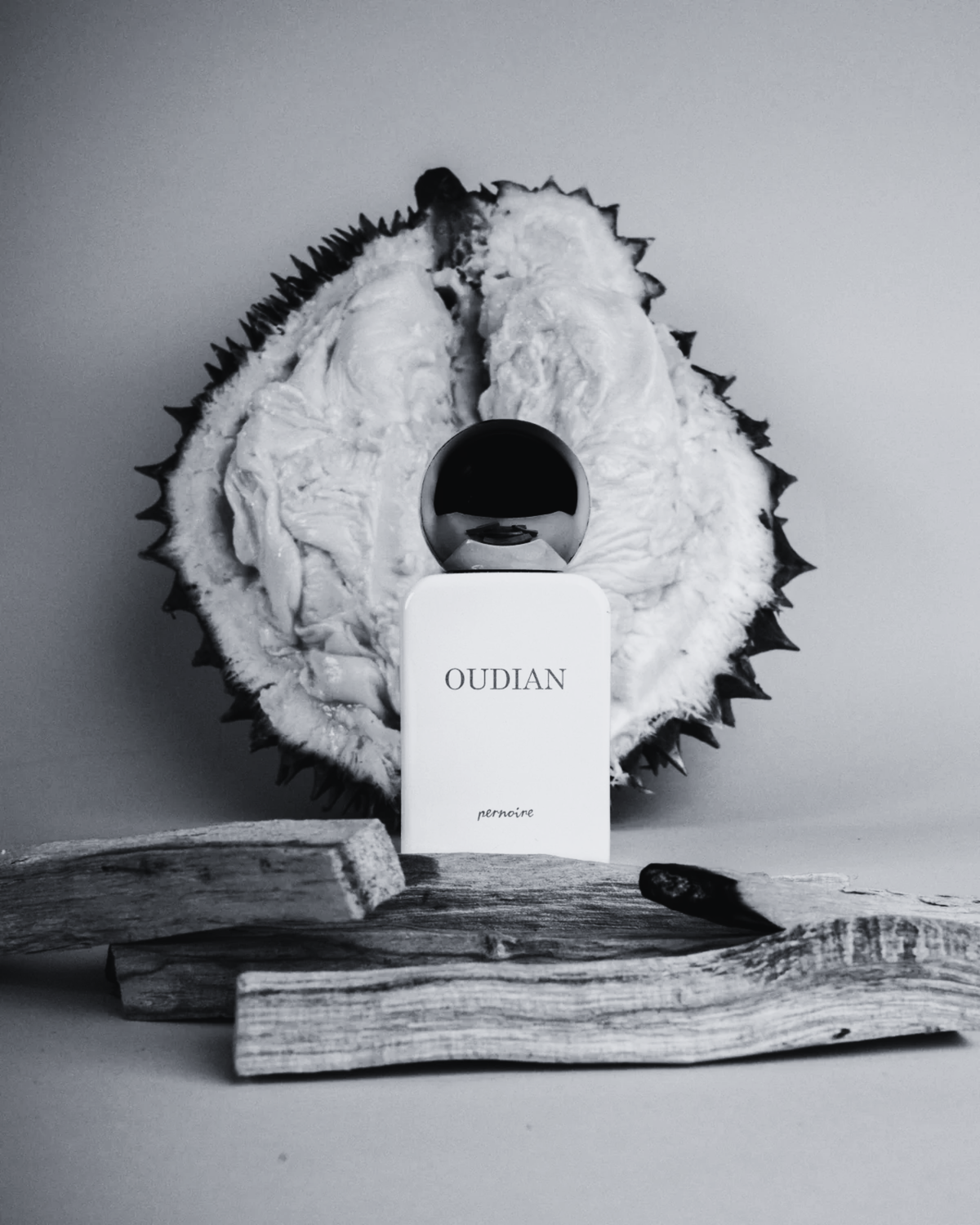 Weisser OUDIAN-Flakon vor aufgeschnittener Durian mit Holzstücken auf hellem Grund.