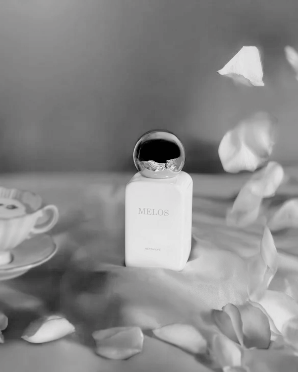 Weisser MELOS-Flakon auf hellem, gefaltetem Stoff neben einer Teetasse und verstreuten Rosenblättern.