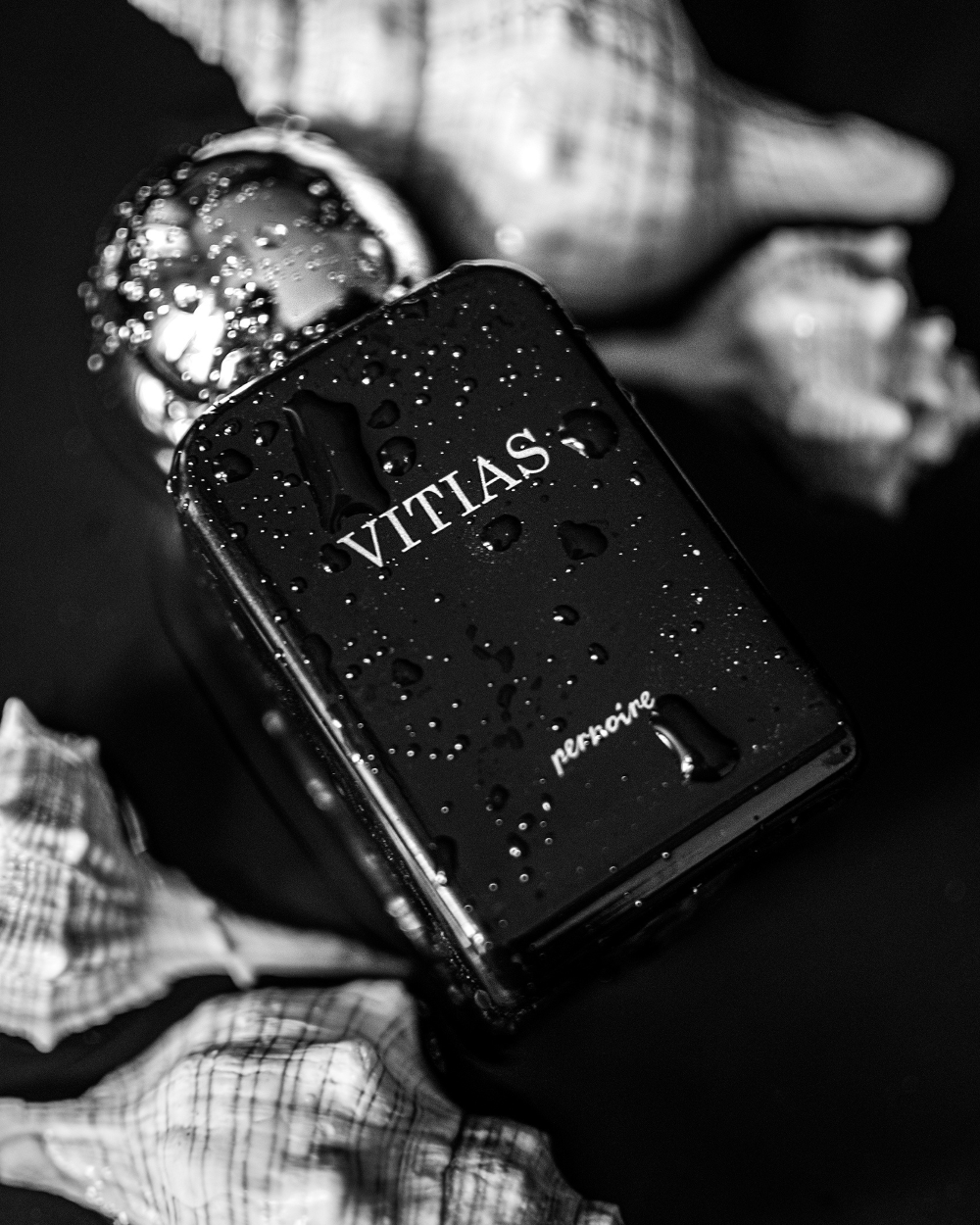 Black VITIAS perfume bottle by pernoire with water droplets, lying beside seashells.