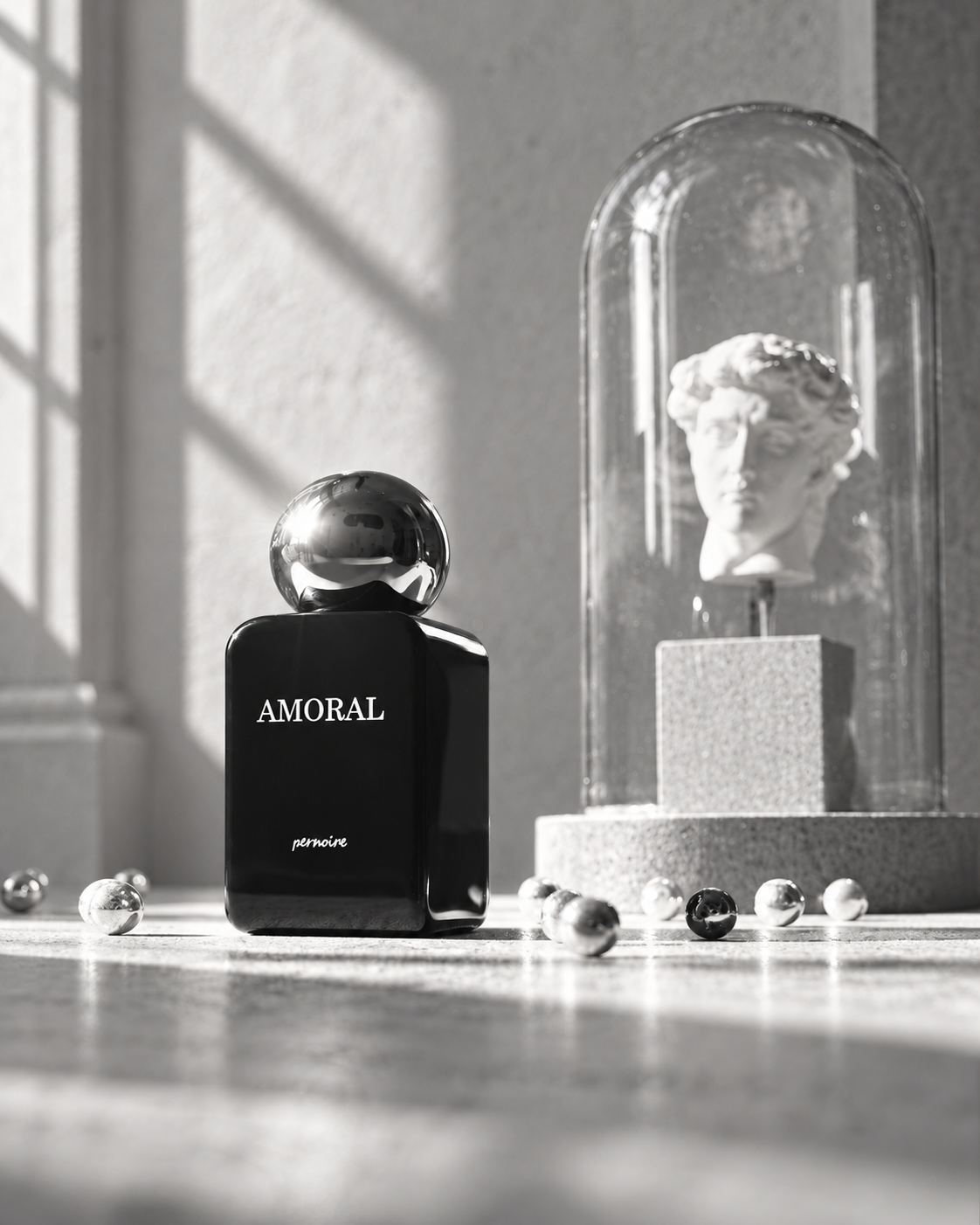 Black AMORAL bottle on pale stone beside a classical head under a glass dome, with window shadows on the wall.