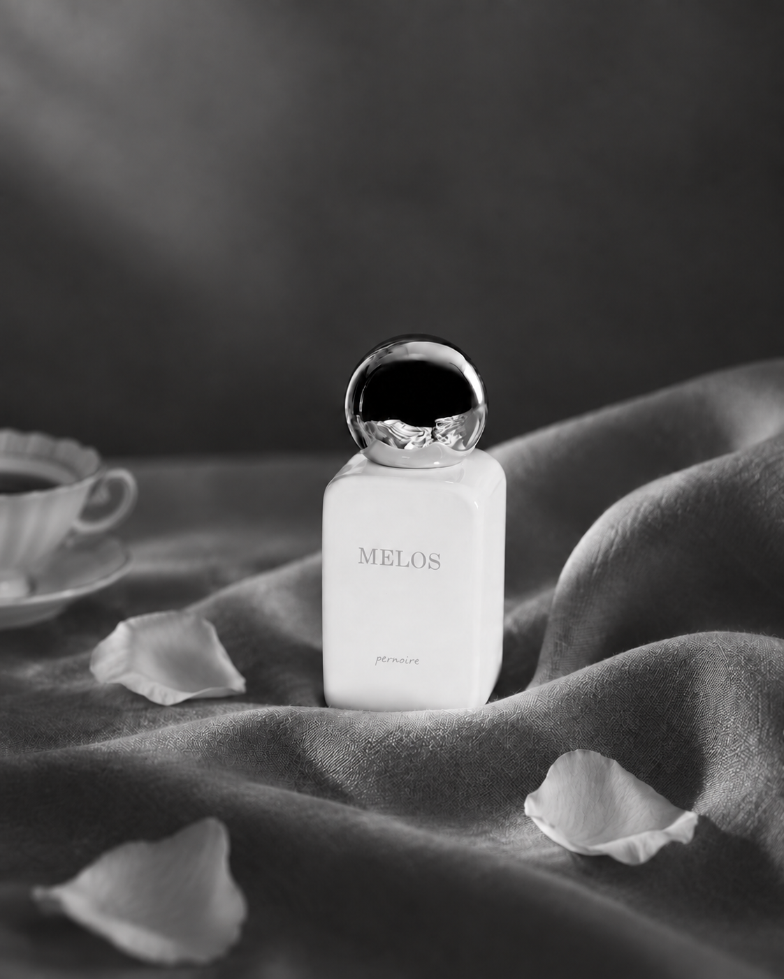 Weisser MELOS-Flakon auf dunklem, gefaltetem Stoff neben einer Teetasse und verstreuten Rosenblättern.